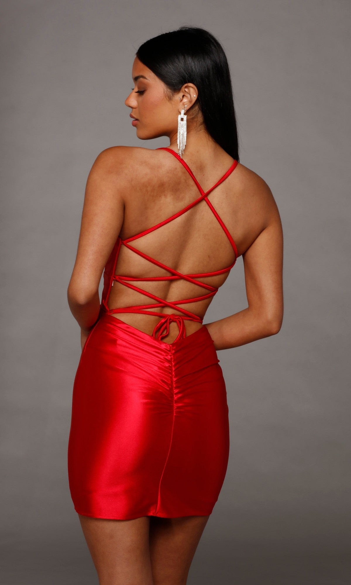 Woman wearing the Lace-Up Short Satin Sheath Hoco Dress: Alyce 40123, featuring crisscross straps and an open back, standing against a plain gray background.